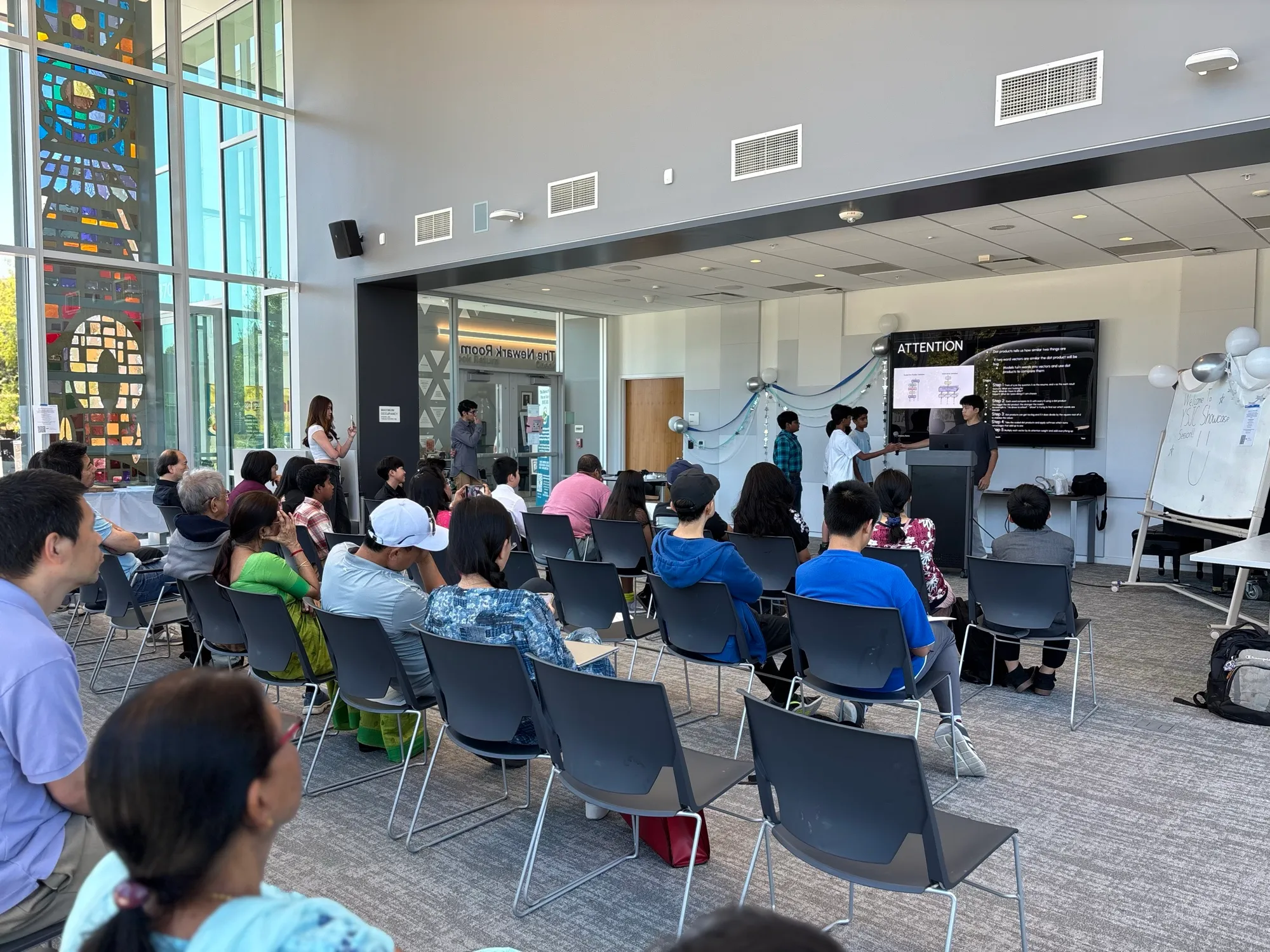 Wide view of the Newark Library Community Room — families seated, CS cohort presenting Attention Is All You Need.